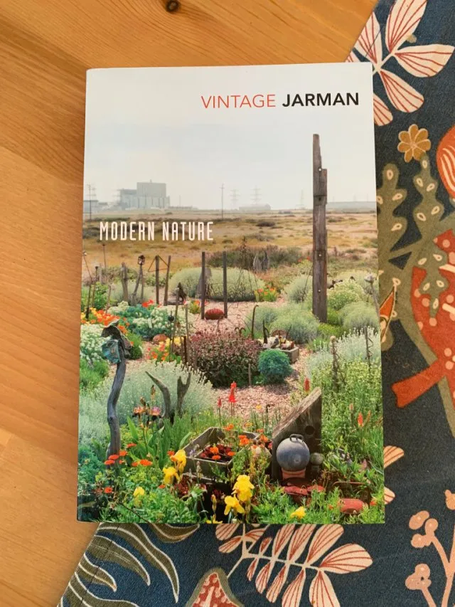Fotografía de la portada del libro. Una planicie llena de malas hierbas con la silueta de una central nuclear al fondo. Más cerca de la cámara, flores de muchos colores pueblan las inmediaciones de la casa de Derek Jarman.