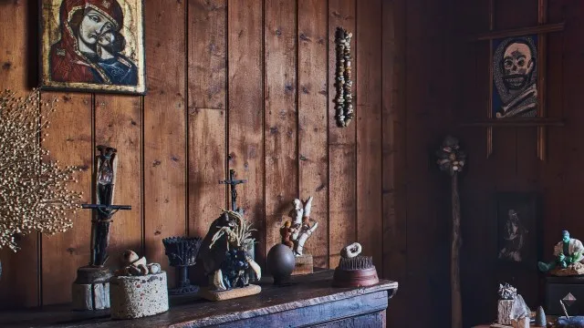 Una pared del interior de la cabaña, hecha de tablas verticales de madera oscura. Hay un cuadro colgado que parece ser un icono de la virgen María acompañando al niño Jesús, de estilo eslavo. Sobre un mueble, pequeñas figuras de madera, cuerda, piedras y ramitas sin formas definidas. A la derecha se ve una figura del increíble Hulk y un mineral sobre un pequeño estandarte, como de una colección.